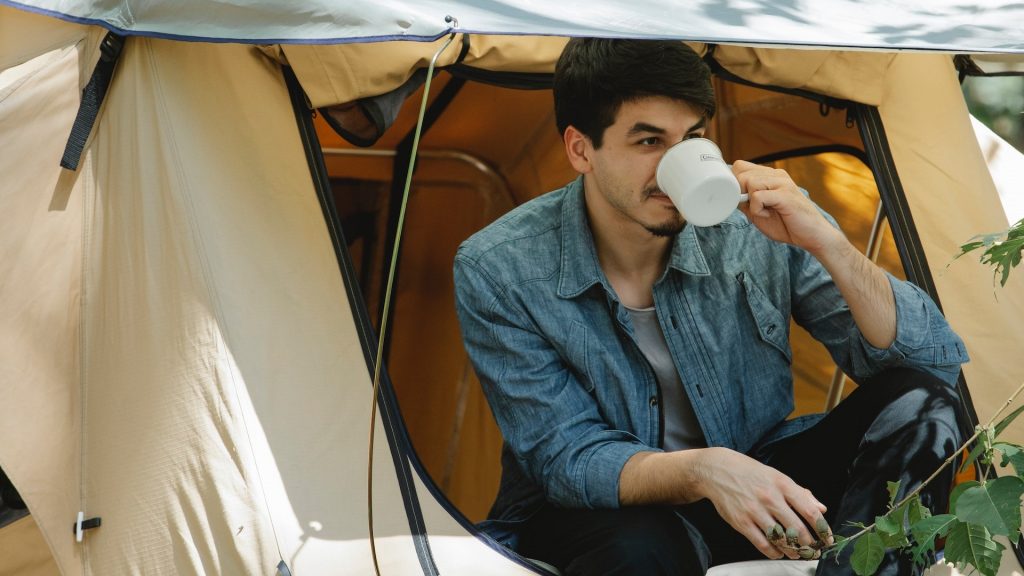Man drinking coffee from a travel mug while sitting at a tent on the roof of a car (credit: Uriel Mont)